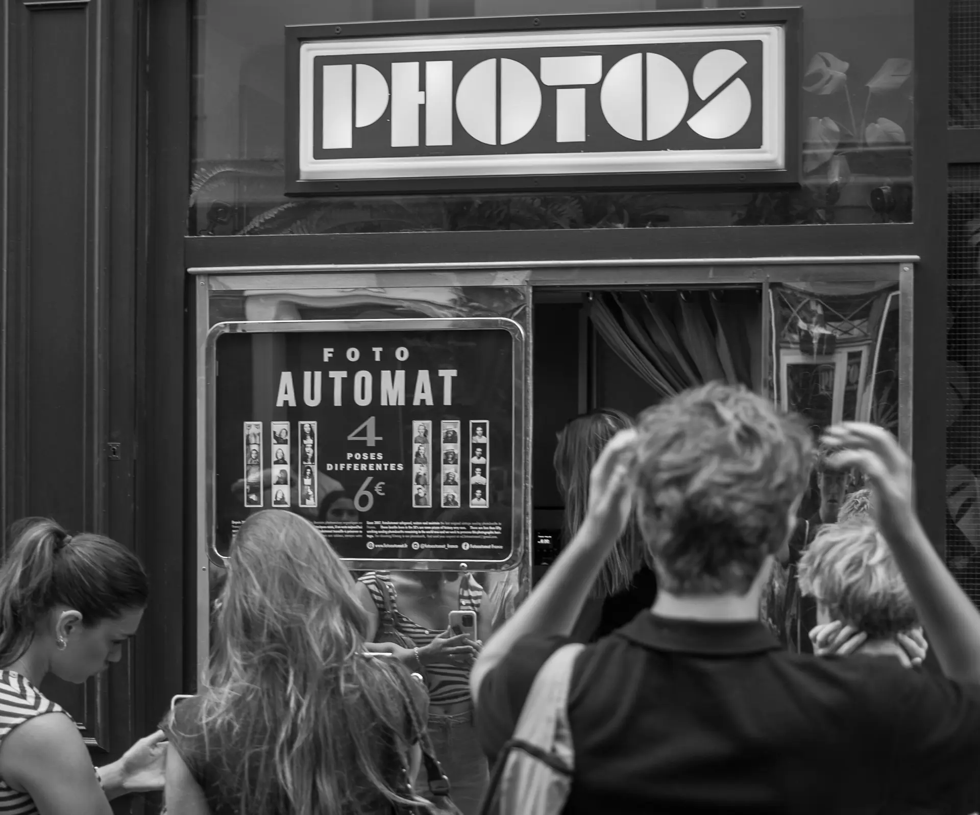 Schwarzweiß-Straßenfoto: Menschen vor einem Fotoautomaten mit Leuchtschild PHOTOS